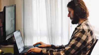 A man sits at a desk in his home, working on his laptop with headphones on