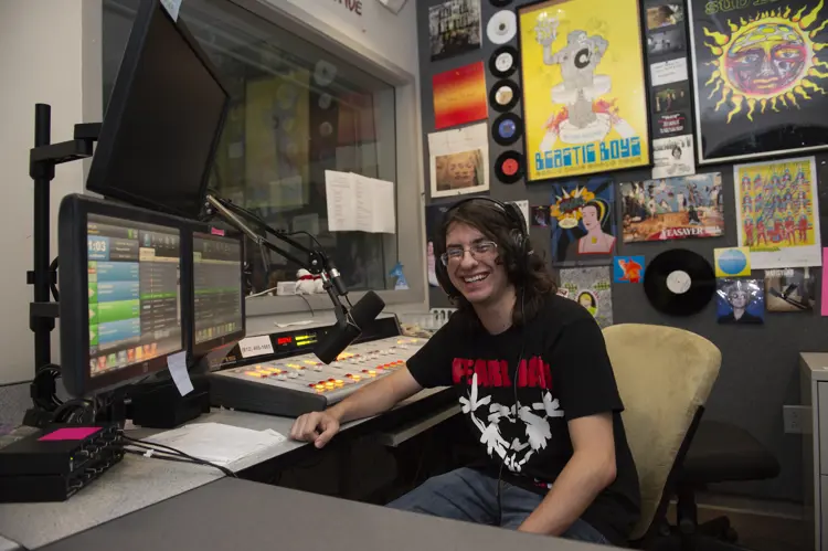 Student DJ smiles from behind the soundboard at the radio studio, which is covered in music posters