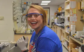 Female medical sciences student smiles while conducting a lab experiment with pipettes, latex gloves, and safety goggles