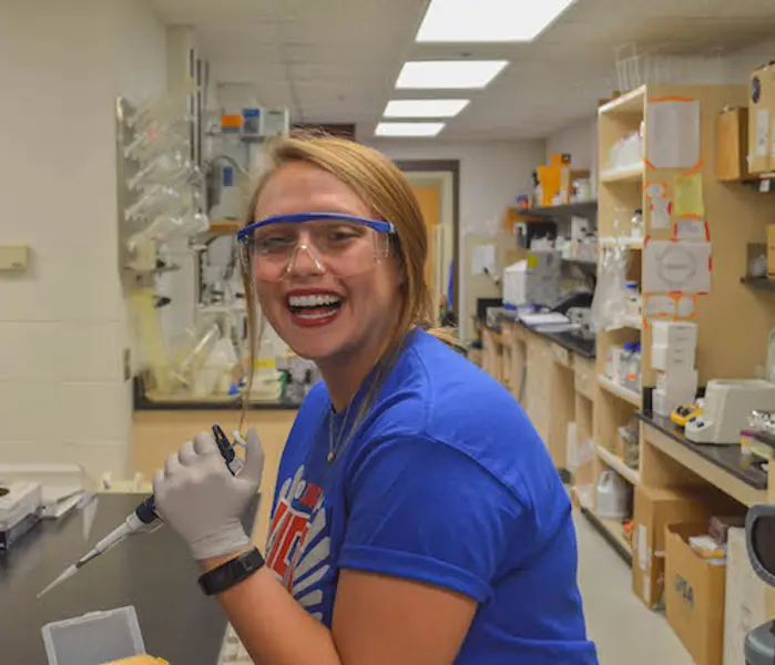 Female medical sciences student smiles while conducting a lab experiment with pipettes, latex gloves, and safety goggles