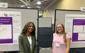 Two USI students in professional attire pose in front of their posters at a conference