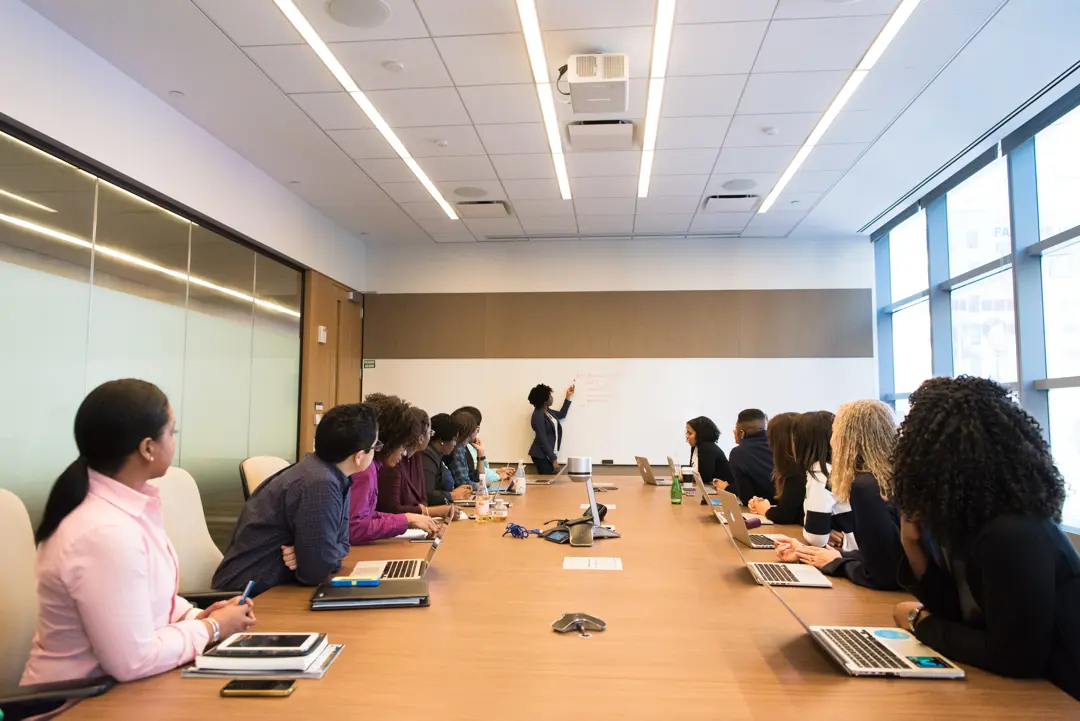An instructor writes on the whiteboard while a group of professionals seated around a conference table observe