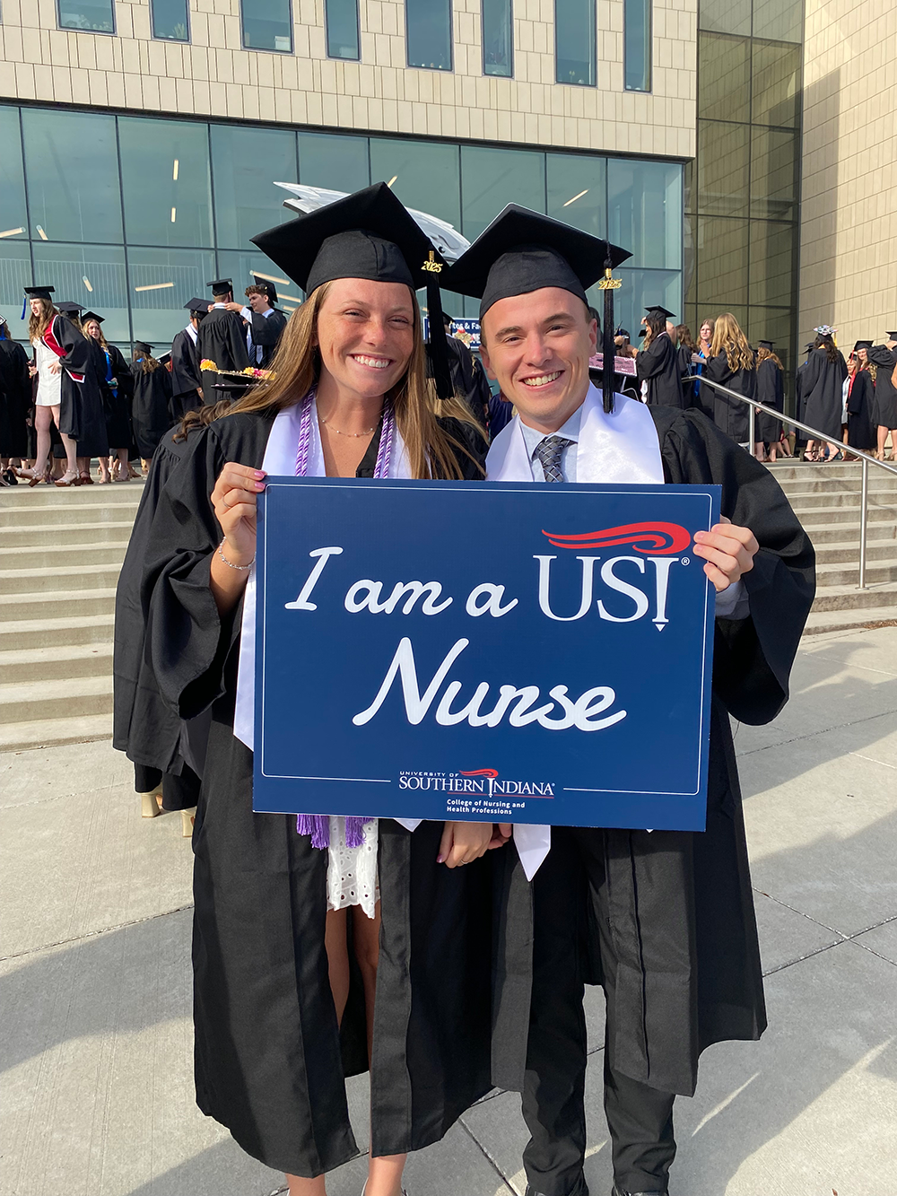 Two nursing students in caps and gowns holding "I am a USI Nurse" sign