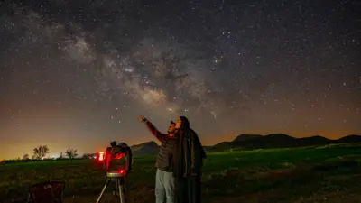 Two people gaze at the starry night sky, one pointing upward