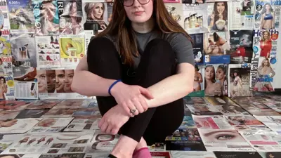 Artist sitting on the floor of an exhibition room. Fashion pages and artwork completely cover the walls and floor.
