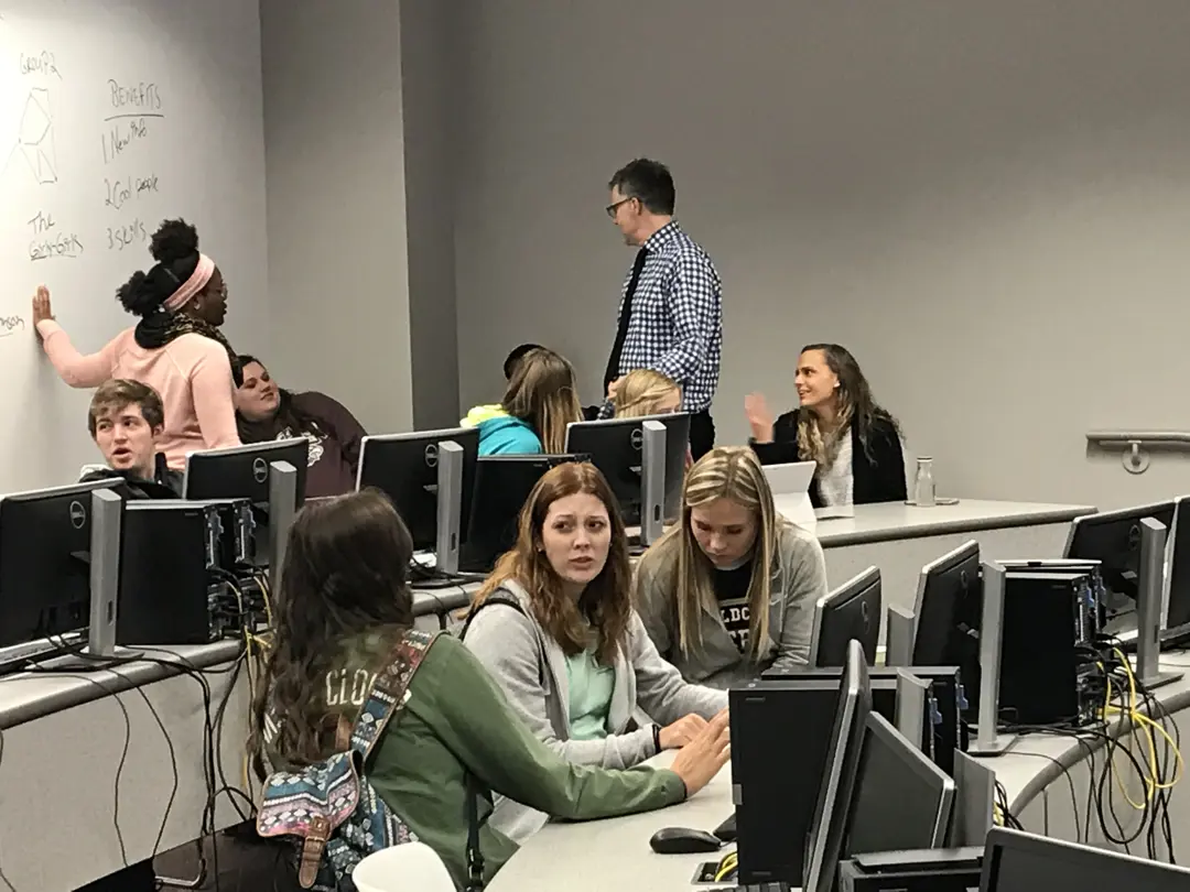Sociology students have a discussion in a computer lab, while one student writes on a whiteboard