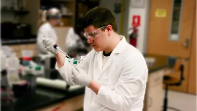 Male student in a white lab coat, latex gloves, and safety glass uses a pipette in the genetics lab