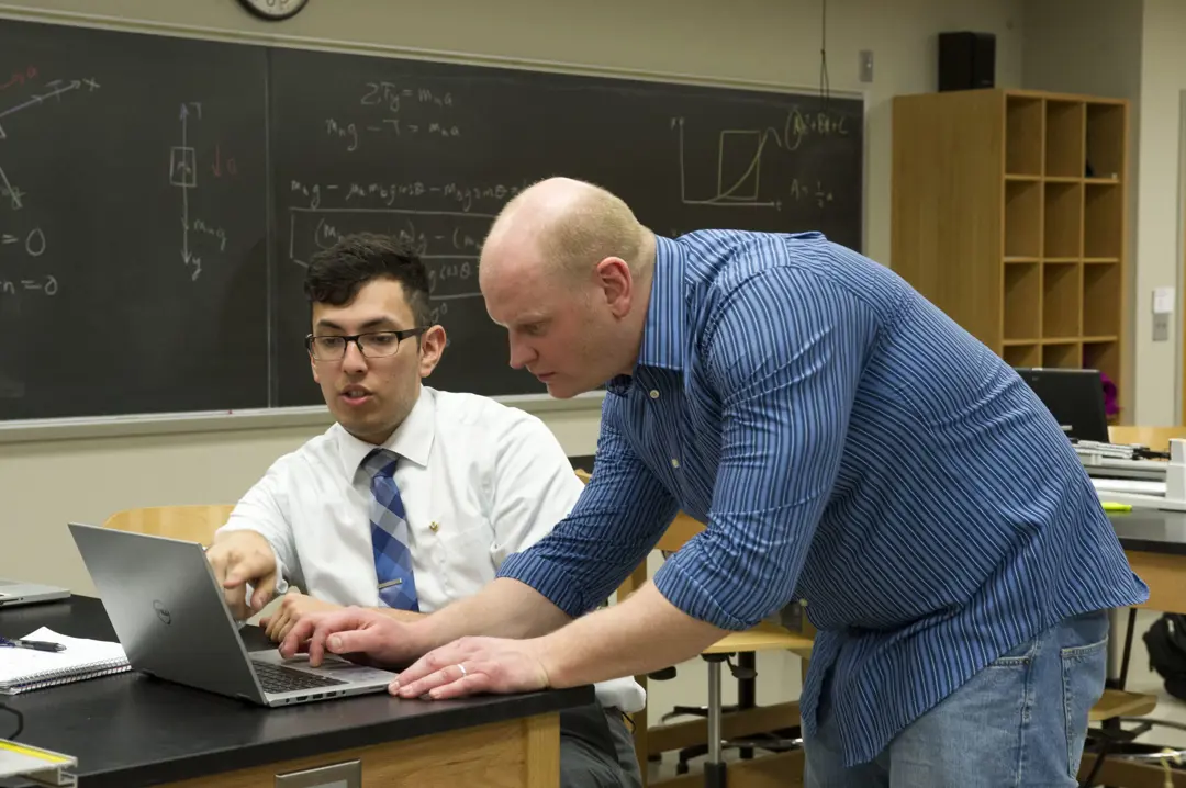 Student and faculty work together on a project on a laptop with a chalkboard in the background with physics equations